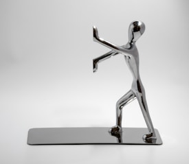 A chrome-plated sculpture of a human figure in a dynamic pose, with one foot forward and arms raised, appearing to push against an invisible barrier. The sculpture is highly polished and reflective, set on a matching rectangular base, all against a plain white background.