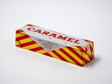 A wrapped caramel wafer biscuit with a silver and red packaging design. Bright yellow and red stripes cover the sides, and text on the top indicates that it is made by T. Tunnock Ltd in Uddingston, Scotland. The packaging includes a weight label of 30g.