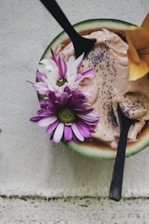 A wooden bowl contains a creamy dessert topped with chia seeds and adorned with colorful flowers. Two wooden spoons are inserted into the dish, creating an inviting presentation. The backdrop is a textured fabric, adding a rustic and natural touch.