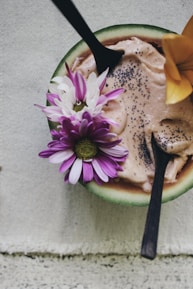 A wooden bowl contains a creamy dessert topped with chia seeds and adorned with colorful flowers. Two wooden spoons are inserted into the dish, creating an inviting presentation. The backdrop is a textured fabric, adding a rustic and natural touch.