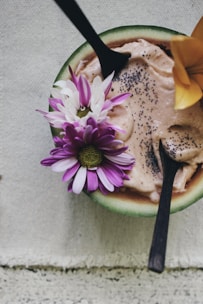A wooden bowl contains a creamy dessert topped with chia seeds and adorned with colorful flowers. Two wooden spoons are inserted into the dish, creating an inviting presentation. The backdrop is a textured fabric, adding a rustic and natural touch.