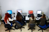Four people are seated at a table, each working on a desktop computer in an office setting. The table has divided workstations, and the people appear engaged with their screens. Various computer peripherals like keyboards and mice are on the table, and a plant is visible in the corner.