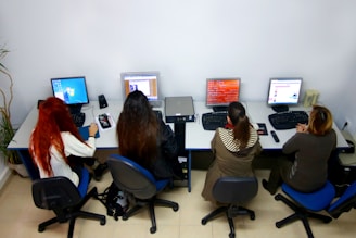 Four people are seated at a table, each working on a desktop computer in an office setting. The table has divided workstations, and the people appear engaged with their screens. Various computer peripherals like keyboards and mice are on the table, and a plant is visible in the corner.