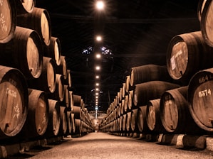 A dimly lit warehouse with rows of large wooden barrels stacked on either side of a central aisle. The barrels are marked with numbers, suggesting storage or aging. Overhead lights create a warm, inviting glow that accentuates the wooden textures and shadows.