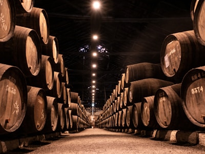 A dimly lit warehouse with rows of large wooden barrels stacked on either side of a central aisle. The barrels are marked with numbers, suggesting storage or aging. Overhead lights create a warm, inviting glow that accentuates the wooden textures and shadows.