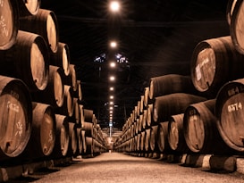 A dimly lit warehouse with rows of large wooden barrels stacked on either side of a central aisle. The barrels are marked with numbers, suggesting storage or aging. Overhead lights create a warm, inviting glow that accentuates the wooden textures and shadows.