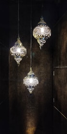 Three intricately designed hanging lamps emit a warm glow. The lamps feature elaborate patterns that create a play of light and shadows on the surrounding dark walls, enhancing their ornate appearance.