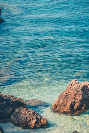 Clear, turquoise water gently laps against rugged rocks along the shoreline, creating a serene and picturesque coastal scene.