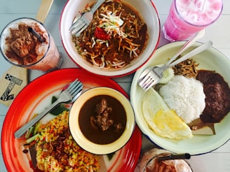 An array of food dishes arranged on a table includes a bowl of noodles with vegetables and meat, a plate of fried rice with vegetables, and another plate containing rice, fried egg, sambal, and anchovies. Beside these dishes are two beverages—one with a pink hue and another with chocolate and whipped cream topping.