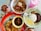 An array of food dishes arranged on a table includes a bowl of noodles with vegetables and meat, a plate of fried rice with vegetables, and another plate containing rice, fried egg, sambal, and anchovies. Beside these dishes are two beverages—one with a pink hue and another with chocolate and whipped cream topping.