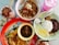An array of food dishes arranged on a table includes a bowl of noodles with vegetables and meat, a plate of fried rice with vegetables, and another plate containing rice, fried egg, sambal, and anchovies. Beside these dishes are two beverages—one with a pink hue and another with chocolate and whipped cream topping.