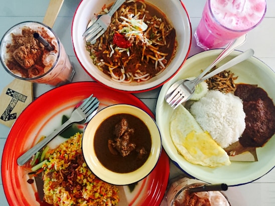 An array of food dishes arranged on a table includes a bowl of noodles with vegetables and meat, a plate of fried rice with vegetables, and another plate containing rice, fried egg, sambal, and anchovies. Beside these dishes are two beverages—one with a pink hue and another with chocolate and whipped cream topping.