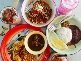 An array of food dishes arranged on a table includes a bowl of noodles with vegetables and meat, a plate of fried rice with vegetables, and another plate containing rice, fried egg, sambal, and anchovies. Beside these dishes are two beverages&mdash;one with a pink hue and another with chocolate and whipped cream topping.