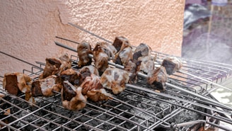 Chunks of meat skewered on metal sticks are placed over a grill, with visible smoke rising from the coals below. The meat appears to be marinated or coated, possibly with a seasoning mixture, highlighting a barbecue setup.
