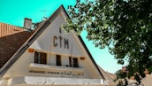 A triangular-roofed building with a sign displaying 'CTM' at the top, surrounded by lush green trees. The lower part of the building features windows and another sign written in French and Arabic. The roof is covered with brown tiles, and the sky is clear and blue.