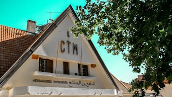 A triangular-roofed building with a sign displaying 'CTM' at the top, surrounded by lush green trees. The lower part of the building features windows and another sign written in French and Arabic. The roof is covered with brown tiles, and the sky is clear and blue.