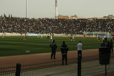 A crowded soccer stadium with a large number of spectators in the stands watching a match. Players on the field are actively involved in the game, wearing contrasting uniforms. The boundary of the field is marked, and there are people standing along the sidelines. The surroundings suggest it is an outdoor venue with urban structures in the background.