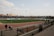 An outdoor sports stadium filled with spectators watching a soccer game. The field is green and well-maintained, surrounded by a track. The stands are packed with people, and there are players and officials on the field. The weather is clear with some clouds.
