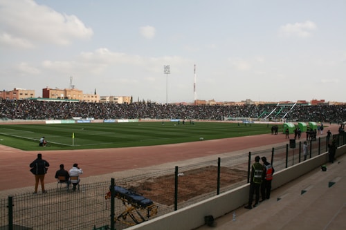 An outdoor sports stadium filled with spectators watching a soccer game. The field is green and well-maintained, surrounded by a track. The stands are packed with people, and there are players and officials on the field. The weather is clear with some clouds.