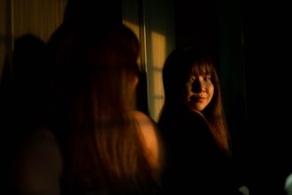 A woman with long hair is gazing at her own reflection in a dimly lit room, with shadows and soft light adding a dramatic effect. The warm ambient light highlights her facial features.