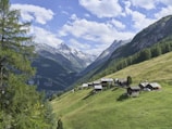 A picturesque alpine scene featuring wooden cabins nestled on a lush green hillside. Majestic snow-capped mountains rise in the background under a partially cloudy sky.