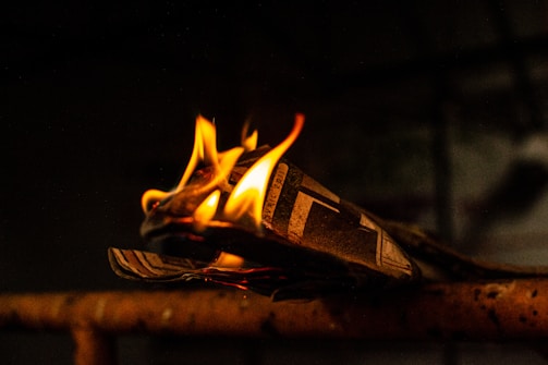A piece of newspaper is partially engulfed in flames, with the bright fire contrasting against a dark background. The flames are vividly yellow and orange, highlighting the texture of the paper as it burns.