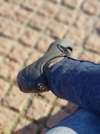 A person is wearing dark shoes with a distinctive branding icon on the side and blue jeans while sitting on a surface made of light brown paving stones.