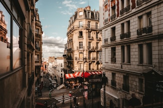 A picturesque urban street scene featuring classic Parisian architecture with ornate balconies. The street is lined with tall buildings, including a hotel, and at the corner, a caf&eacute; with a red and white striped awning. Pedestrians and street signs add to the bustling city atmosphere.