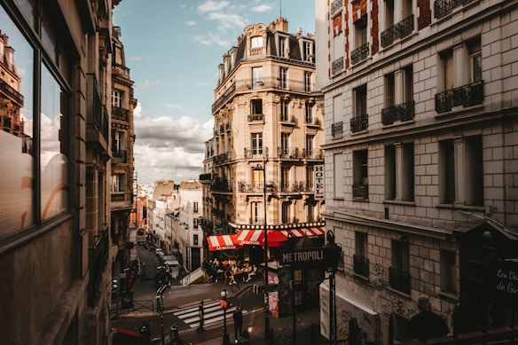 A picturesque urban street scene featuring classic Parisian architecture with ornate balconies. The street is lined with tall buildings, including a hotel, and at the corner, a café with a red and white striped awning. Pedestrians and street signs add to the bustling city atmosphere.