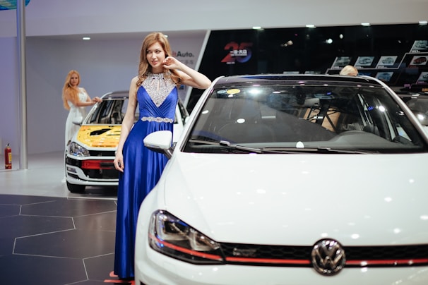 A woman in a blue, elegant dress poses beside a white Volkswagen car in a showroom. Another woman in a white dress interacts with a nearby car which has a yellow hood. The setting features various cars and a dark wall showcasing car images in the background.