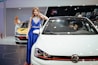 A woman in a blue, elegant dress poses beside a white Volkswagen car in a showroom. Another woman in a white dress interacts with a nearby car which has a yellow hood. The setting features various cars and a dark wall showcasing car images in the background.