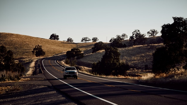 A winding road stretches across a rural landscape with dry grassy hills flanking either side. Sparse trees are scattered across the hills, casting shadows as the sun sets, creating a tranquil and serene ambiance. A vintage car is driving down the road, heading away into the distance.