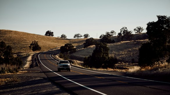 A winding road stretches across a rural landscape with dry grassy hills flanking either side. Sparse trees are scattered across the hills, casting shadows as the sun sets, creating a tranquil and serene ambiance. A vintage car is driving down the road, heading away into the distance.