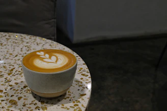 A warm latte in a cream-colored cup on a wooden table with soft blush and terracotta accents around.