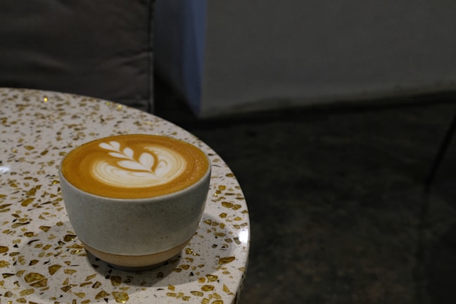 A warm latte with intricate foam art resting on a black charcoal table with gold accents.