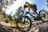 A person wearing a bright yellow and black cycling outfit is riding a mountain bike on a rocky terrain surrounded by lush green trees under a clear blue sky. The image captures the action and intensity of cycling in a natural setting.