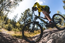 A person wearing a bright yellow and black cycling outfit is riding a mountain bike on a rocky terrain surrounded by lush green trees under a clear blue sky. The image captures the action and intensity of cycling in a natural setting.