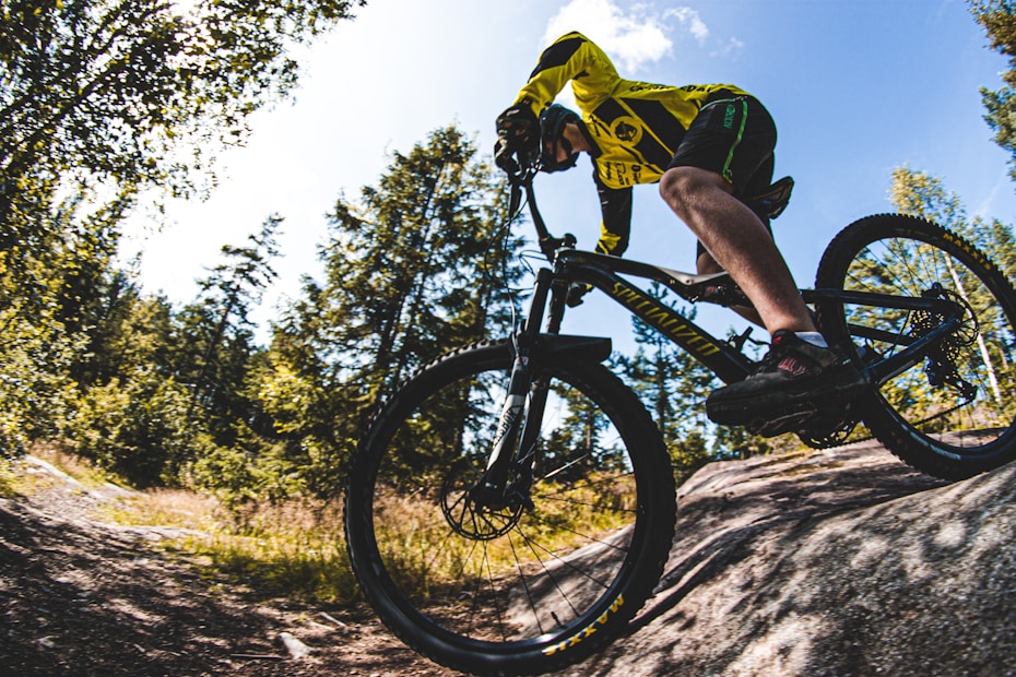 A person wearing a bright yellow and black cycling outfit is riding a mountain bike on a rocky terrain surrounded by lush green trees under a clear blue sky. The image captures the action and intensity of cycling in a natural setting.