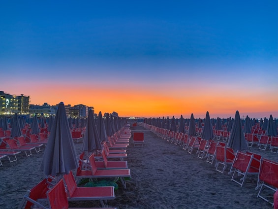 Rows of red beach chairs and closed umbrellas are lined up symmetrically on a sandy beach with buildings visible in the background. The sky is a vibrant gradient of blue transitioning to a warm orange near the horizon, indicating either sunrise or sunset.
