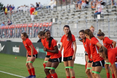 A group of female soccer players is engaged in a warm-up session on a grass field. They are dressed in red shirts with black shorts, and some of them are wearing green resistance bands around their thighs. Behind them, bleachers filled with spectators are visible, suggesting a sports event atmosphere.