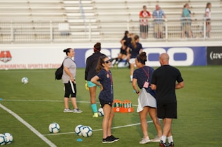A group of people are gathered on a soccer field, likely during a training session. Some individuals are wearing athletic gear and appear to be coaching or instructing, while others are moving in the background. There are soccer balls scattered on the grass, and orange cones mark certain areas. A nearby bench is visible with some seated spectators.