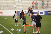 A group of people are gathered on a soccer field, likely during a training session. Some individuals are wearing athletic gear and appear to be coaching or instructing, while others are moving in the background. There are soccer balls scattered on the grass, and orange cones mark certain areas. A nearby bench is visible with some seated spectators.