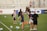 A group of people are gathered on a soccer field, likely during a training session. Some individuals are wearing athletic gear and appear to be coaching or instructing, while others are moving in the background. There are soccer balls scattered on the grass, and orange cones mark certain areas. A nearby bench is visible with some seated spectators.