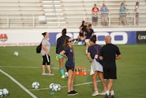 A group of people are gathered on a soccer field, likely during a training session. Some individuals are wearing athletic gear and appear to be coaching or instructing, while others are moving in the background. There are soccer balls scattered on the grass, and orange cones mark certain areas. A nearby bench is visible with some seated spectators.