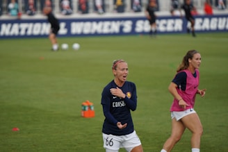 Two female soccer players are on a grassy field, engaged in a soccer practice or game. One player in a navy shirt appears to be in action, possibly giving instructions or reacting to the game. Another in a pink bib runs nearby. The background shows a banner for a women's soccer league and several blurred players and spectators.