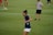 A person in a navy blue sports shirt and white shorts is actively engaged in a sporting event on a grassy field. The background shows other individuals, also dressed in sports attire, possibly participating in a group activity or practice session.