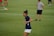 A person in a navy blue sports shirt and white shorts is actively engaged in a sporting event on a grassy field. The background shows other individuals, also dressed in sports attire, possibly participating in a group activity or practice session.