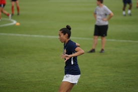 A person in a navy blue sports shirt and white shorts is actively engaged in a sporting event on a grassy field. The background shows other individuals, also dressed in sports attire, possibly participating in a group activity or practice session.