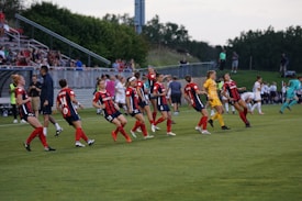 A group of female soccer players wearing red and navy blue uniforms are seen on a soccer field. They appear to be warming up or performing a choreographed routine, with some players in stride and others standing still. A player in a yellow goalkeeper uniform is also present. In the background, there are spectators seated on bleachers, and more players are visible further along the field.