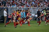 A group of female soccer players in red and blue uniforms are actively running towards the field, indicating a substitution or start of the game. The scene is vibrant with many spectators in the background, sitting on bleachers and watching the event. There are coaching staff and team members on the sidelines, along with sports equipment such as water coolers.